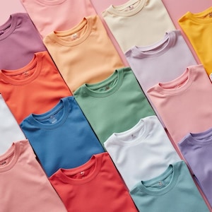 Könnte beinhalten: Eine flache Anordnung von 14 einfarbigen T-Shirts in verschiedenen Farben, darunter Rosa, Orange, Grün, Blau, Weiß und Lila. Die Shirts sind in einem versetzten Muster auf einem rosa Hintergrund angeordnet.