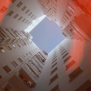K&ouml;nnte beinhalten: Eine Ansicht, die vom Himmel aus einer engen Gasse zwischen zwei hohen wei&szlig;en Geb&auml;uden nach oben blickt. Die Geb&auml;ude haben viele Fenster und Balkone. Der Himmel ist strahlend blau und es gibt einen roten Schein von der Sonne.