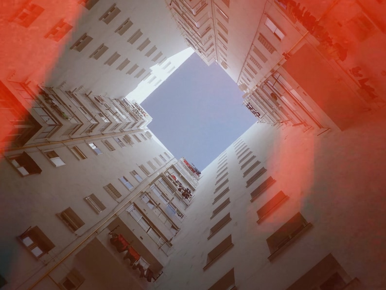 K&ouml;nnte beinhalten: Eine Ansicht, die vom Himmel aus einer engen Gasse zwischen zwei hohen wei&szlig;en Geb&auml;uden nach oben blickt. Die Geb&auml;ude haben viele Fenster und Balkone. Der Himmel ist strahlend blau und es gibt einen roten Schein von der Sonne.