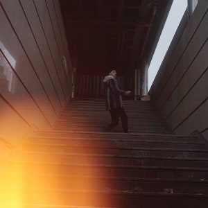 K&ouml;nnte beinhalten: Eine Person geht eine Betontreppe in einem schwach beleuchteten Flur hinauf. Die Treppe ist aus Beton und hat eine abgenutzte, strukturierte Oberfl&auml;che. Der Flur ist schmal und hat eine Betondecke.