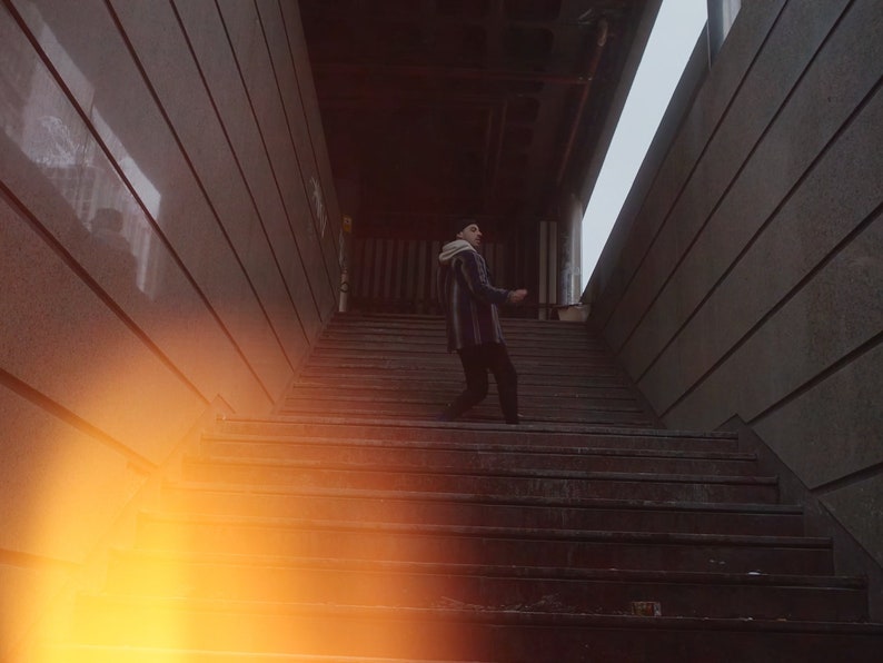 K&ouml;nnte beinhalten: Eine Person geht eine Betontreppe in einem schwach beleuchteten Flur hinauf. Die Treppe ist aus Beton und hat eine abgenutzte, strukturierte Oberfl&auml;che. Der Flur ist schmal und hat eine Betondecke.
