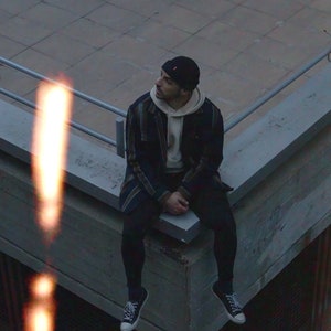 K&ouml;nnte beinhalten: Eine Person mit einer schwarzen M&uuml;tze, einem karierten Hemd, einem wei&szlig;en Hoodie, schwarzen Hosen und schwarzen Sneakern sitzt auf einer Betonkante.
