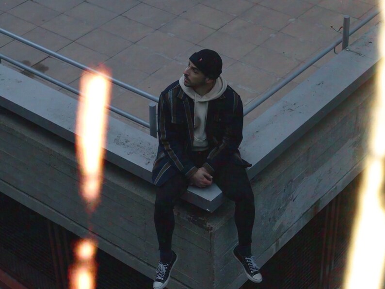 K&ouml;nnte beinhalten: Eine Person mit einer schwarzen M&uuml;tze, einem karierten Hemd, einem wei&szlig;en Hoodie, schwarzen Hosen und schwarzen Sneakern sitzt auf einer Betonkante.