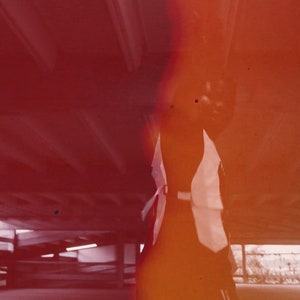 K&ouml;nnte beinhalten: Ein verschwommenes Bild einer Person, die in einer Tiefgarage steht. Die Person tr&auml;gt ein wei&szlig;es Hemd und eine schwarze Hose. Das Bild ist unscharf und hat einen roten Farbton.