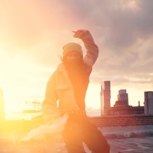 K&ouml;nnte beinhalten: Eine Person in einer gr&uuml;nen M&uuml;tze und einer braunen Jacke posiert auf einem Dach mit einer Skyline im Hintergrund. Die Sonne geht hinter der Person unter und erzeugt einen warmen Schein.
