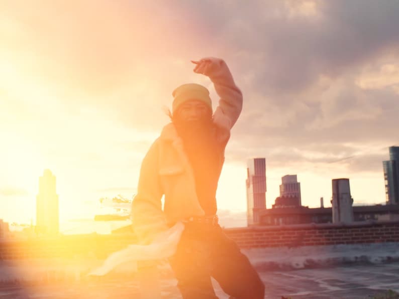 K&ouml;nnte beinhalten: Eine Person in einer gr&uuml;nen M&uuml;tze und einer braunen Jacke posiert auf einem Dach mit einer Skyline im Hintergrund. Die Sonne geht hinter der Person unter und erzeugt einen warmen Schein.