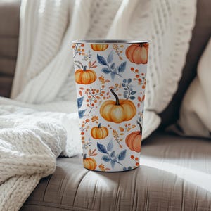 Op de afbeelding: Een witte beker met een patroon van oranje pompoenen, blauwe bladeren en oranje bessen. De cilindrische beker lijkt van roestvrij staal te zijn. De achtergrond toont een gezellige woonkamer met een witte deken.