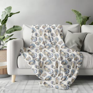 May include: A white throw blanket draped over a sofa, featuring a repeating pattern of watercolor oyster illustrations in shades of blue, beige, and brown. The blanket is made of a soft material and adds a coastal theme to the living room.