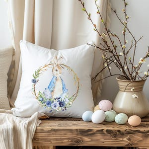 May include: A white pillow with a watercolor illustration of a bunny rabbit wearing a blue jacket and sitting in a wreath of blue flowers. The pillow is on a wooden surface with pastel colored Easter eggs.
