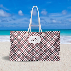 Op de afbeelding: Een rode en zwarte geruite strandtas met witte touwhandvatten. De tas heeft een gepersonaliseerd naamplaatje met de initialen "JAS" in bruine letters.