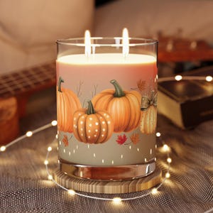 May include: A glass candle with a pumpkin design. The candle has two wicks and is set on a wooden coaster. The candle features orange pumpkins with green stems and fall leaves. The candle is surrounded by string lights.
