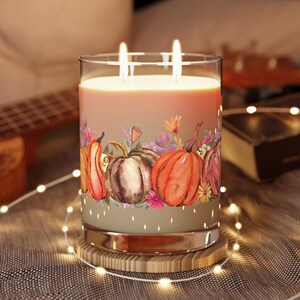May include: A large glass candle with two lit wicks, featuring a watercolor design of pumpkins and flowers. The candle sits on a wooden coaster, surrounded by string lights. The candle's wax has a warm, orange hue.