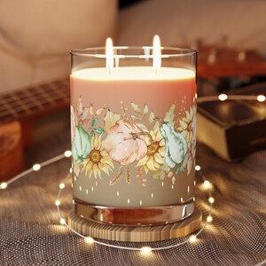 May include: A large glass candle with two lit wicks. The candle has a peach-colored wax and is decorated with a floral design featuring pumpkins, sunflowers, and gourds. It sits on a wooden coaster, with string lights around it.