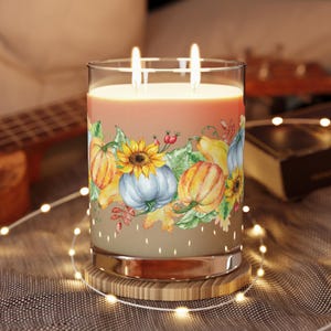 May include: A large glass candle with two wicks, featuring a fall-themed design. The candle has a light orange wax and is decorated with watercolor pumpkins, sunflowers, and leaves. It sits on a wooden coaster, surrounded by fairy lights.