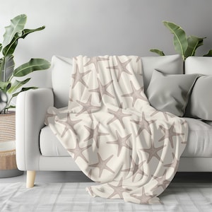 May include: A cream-colored throw blanket draped over a white sofa. The blanket features a repeating pattern of taupe starfish. The sofa is in a living room setting with plants in the background.