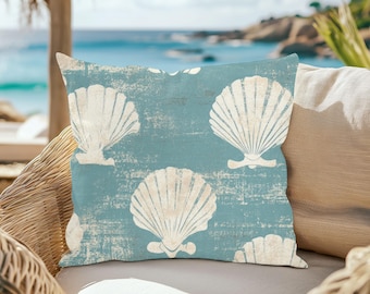 May include: Square throw pillow with a light blue background and a pattern of large, white seashells. The pillow is on a wicker chair with a beach scene in the background.