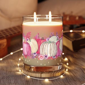 May include: A large glass candle with two wicks, featuring a watercolor design of pumpkins in shades of pink, white, and orange. The candle sits on a wooden coaster, with string lights in the foreground.