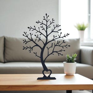 May include: A black metal tree-shaped jewelry organizer with a heart-shaped cutout at the base. The tree has many branches for hanging necklaces and earrings. It is displayed on a wooden table with a small potted plant.