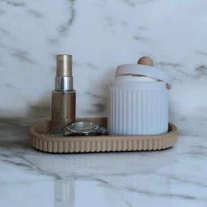 May include: A light brown oval tray with a textured edge, holding a white ceramic container with a lid, a small bottle of liquid, and a silver watch.