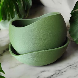 May include: A green ceramic planter with a wavy design and a matching saucer. The planter is made of a textured material and has a smooth, rounded shape.