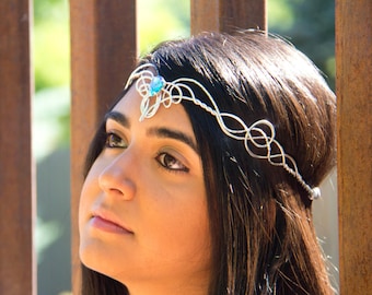 May include: A woman with long dark hair wearing a silver wire headband with a blue gemstone. The headband is adorned with intricate designs.