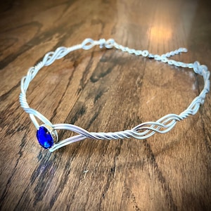 May include: A silver-toned wire crown with a large, oval, blue gemstone at the center. The wire is intricately woven in a braided pattern. The crown is designed to be worn around the head, with a delicate chain for adjustment. The gemstone is a vibrant, deep blue.