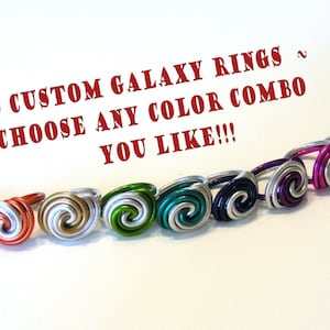 May include: A row of eight custom galaxy rings, each with a different colour combination. The rings are made of wire and have a spiral design. The text "CUSTOM GALAXY RINGS ~ CHOOSE ANY COLOUR COMBO YOU LIKE!!!" is written above the rings.