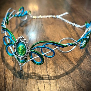 May include: A silver wire headband with green and blue wire accents. The headband has a decorative, ornate, silver setting with a green and blue iridescent gemstone.
