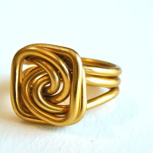 May include: A gold ring with a square-shaped knot design. The knot is made of multiple interwoven strands of metal.