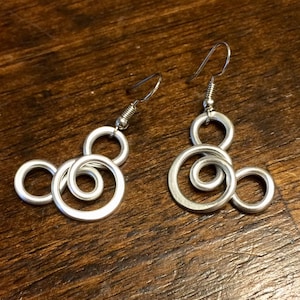 May include: A pair of silver earrings with a unique design featuring three interconnected circles. The earrings are made of metal and have a simple, modern look.