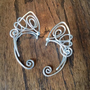 May include: A pair of silver ear cuffs with intricate, swirling designs. The cuffs are made of wire and have a delicate, feminine look.