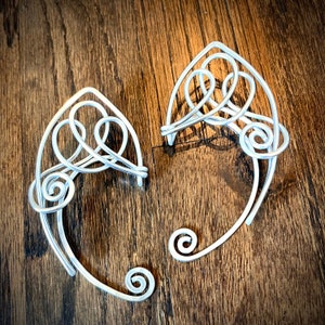 May include: A pair of silver wire ear cuffs with a delicate, intricate design. The cuffs are made of thin wire that is twisted and shaped into a series of loops and swirls.