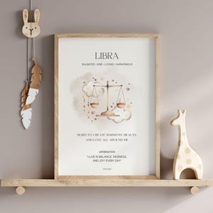May include: A framed Libra zodiac sign print with a beige background. The print features a scale illustration, text that reads "LIBRA", and affirmations. A wooden giraffe and a decorative bunny with feathers are on a shelf below the print.