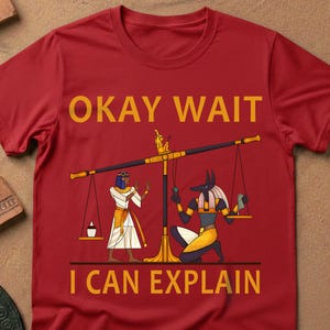 May include: Red t-shirt with the text "OKAY WAIT I CAN EXPLAIN" in gold letters. The shirt features an illustration of Egyptian figures and a balance scale. The design is centered on the front of the shirt.