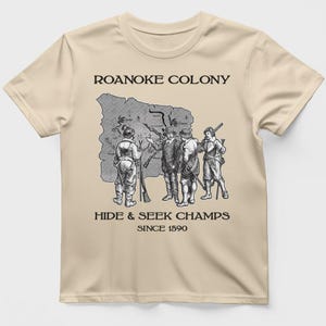 May include: Beige t-shirt featuring a black and white illustration of historical figures gathered around a map. The text "ROANOKE COLONY" is above the illustration, and "HIDE & SEEK CHAMPS SINCE 1590" is below.