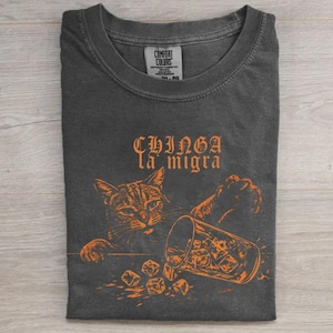 May include: Dark gray t-shirt with an orange graphic of a cat knocking over a glass of ice cubes. The text "CHINGA la migra" is above the cat and glass. The shirt is folded on a light wood surface.