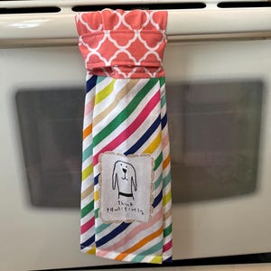 Könnte beinhalten: Ein buntes, gestreiftes Küchentuch mit einem rosa-weißen Muster oben. Das Tuch hat einen weißen Fleck mit einer schwarz-weißen Illustration eines Hundes und dem Text "Think pawsitively".