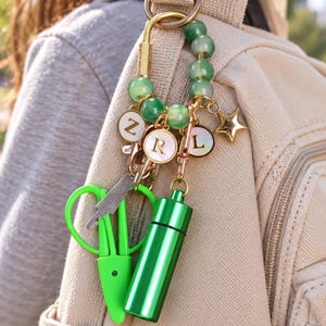 May include: A gold-toned carabiner keychain featuring green marbled beads, a gold star charm, and three circular white charms with the letters Z, R, and L. Attached are a bright green pair of small scissors and a matching green cylindrical metal pill case.
