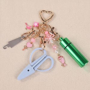 May include: A gold-coloured keyring with various charms. Charms include a small saw, light blue scissors, a green cylinder, and pink and white decorative elements. The keyring has a heart-shaped clasp.