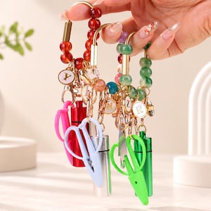 May include: A collection of four beaded keychains featuring gold-toned metal clasps, each attached to a small cylindrical container and a pair of plastic scissors. The beads are red, green, and multi-coloured, with circular charms displaying the letters Y, W, Z, and R.