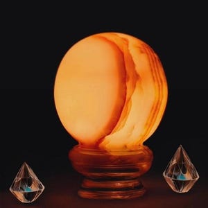 Puede incluir: Una gran lámpara de sal redonda, naranja y blanca con una base de madera. La lámpara está encendida y brilla.