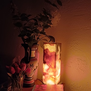 May include: A warm, glowing lamp made of onyx with a marbled pattern. The lamp is on a table with a book and a vase with flowers.