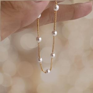 Könnte beinhalten: Eine goldene Kettenkette mit weißen Perlenakzenten. Die Kette ist zart und hat ein schlichtes Design.