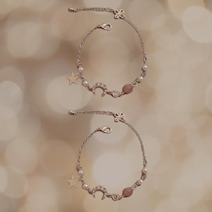 May include: Two delicate gold chain bracelets with star and crescent moon charms. Each bracelet features small white pearls and a pink stone.