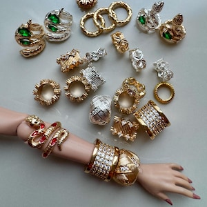 Op de afbeelding: Een assortiment gouden en zilveren ringen en armbanden, sommige versierd met edelstenen. De sieraden omvatten slangenringen met rode accenten en brede gouden armbanden met heldere stenen. Verschillende ringontwerpen zijn zichtbaar.
