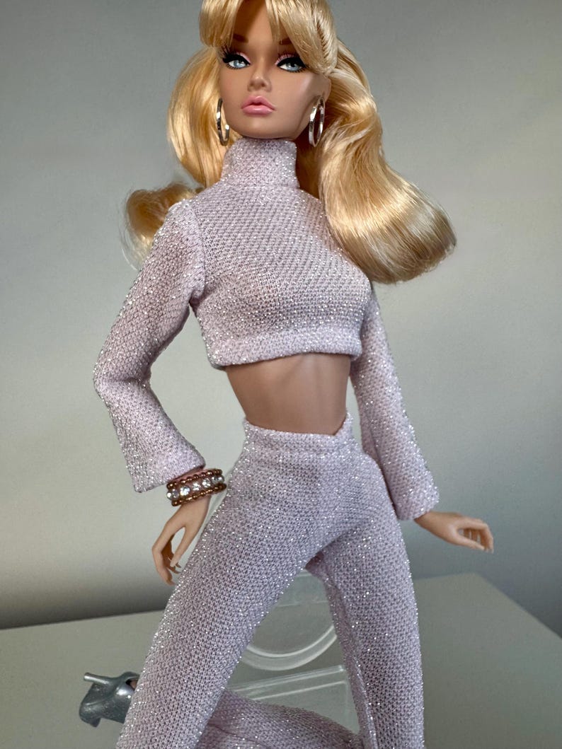 Pu&ograve; includere: Bambola alla moda con capelli biondi e occhi blu, che indossa un completo a due pezzi lilla chiaro e scintillante. L'outfit comprende un top corto a collo alto e pantaloni svasati. La bambola indossa anche orecchini a cerchio e un braccialetto.