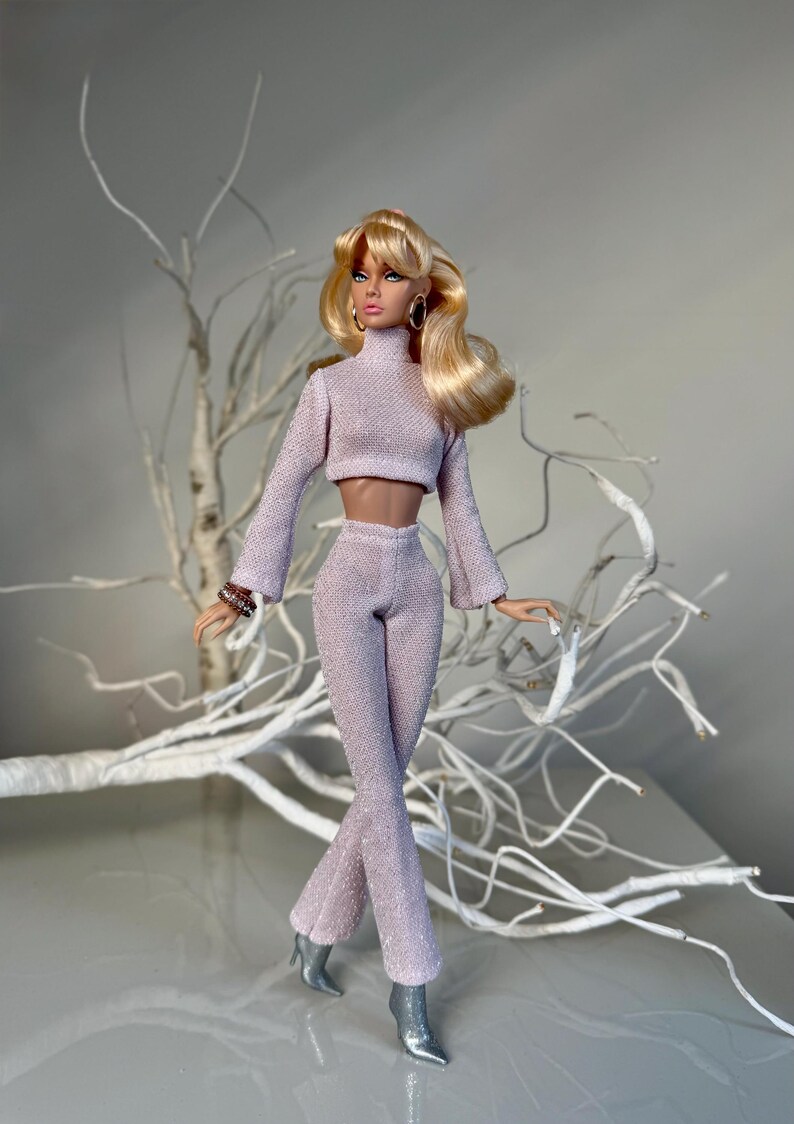 Pu&ograve; includere: Bambola alla moda con un completo a due pezzi strutturato viola chiaro con pantaloni svasati e un top corto. La bambola ha i capelli biondi, orecchini a cerchio dorati e stivali argentati. Un ramo bianco decorativo &egrave; sullo sfondo.
