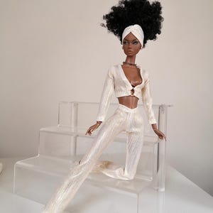 May include: A fashion doll with dark brown skin, black curly hair, and a white turban is wearing a white, sparkly, two-piece outfit. The doll is sitting on a clear acrylic tiered stand. The doll is wearing gold sandals.