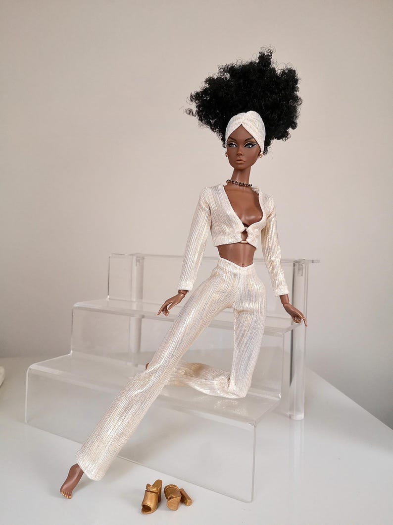 May include: A fashion doll with dark brown skin, black curly hair, and a white turban is wearing a white, sparkly, two-piece outfit. The doll is sitting on a clear acrylic tiered stand. The doll is wearing gold sandals.