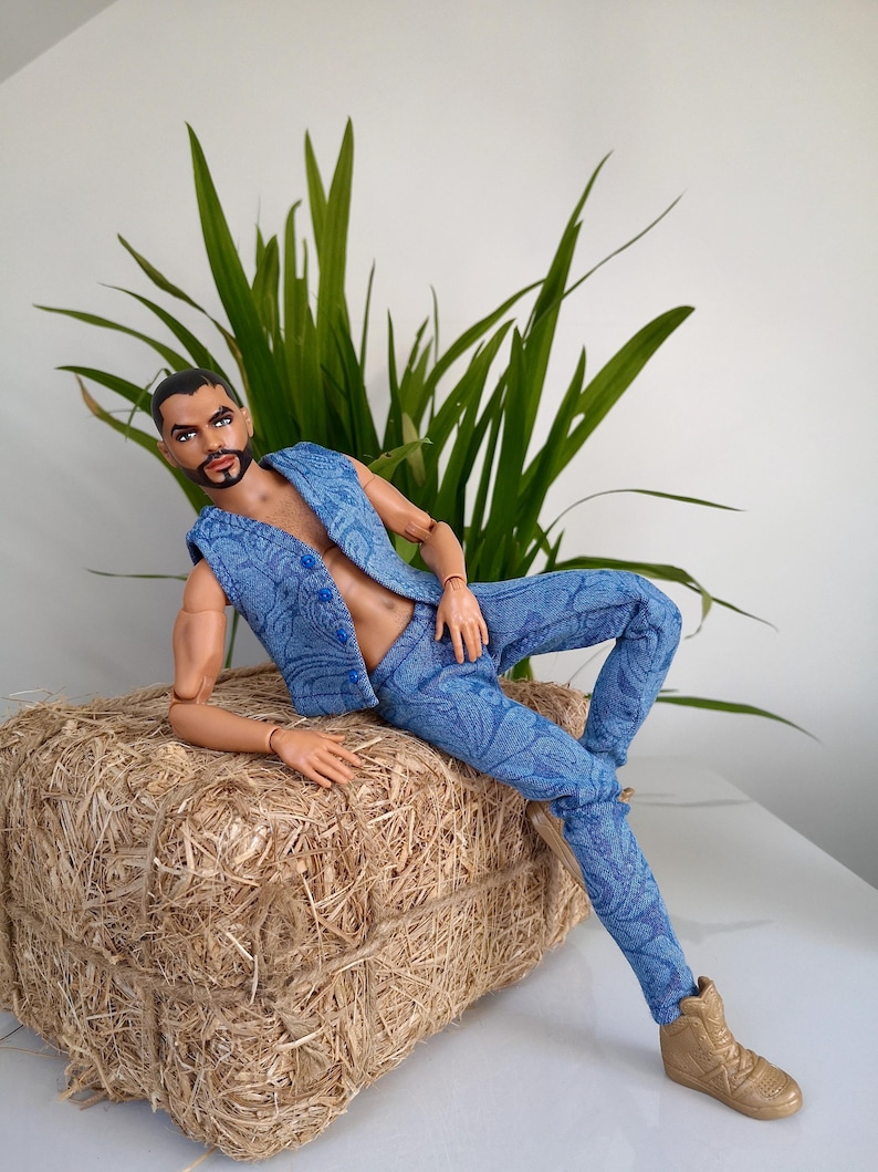 May include: A doll wearing a blue paisley vest and pants, paired with gold sneakers. The doll is posed on a hay bale, with green plants in the background. The doll has a beard and short dark hair.